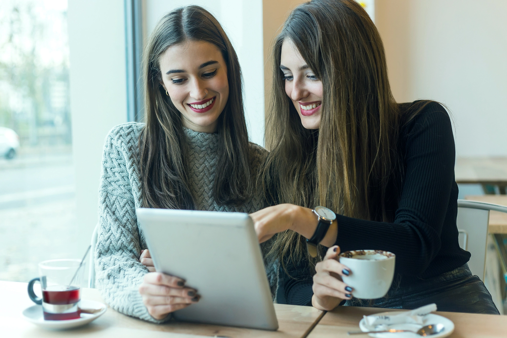 Two smiling young women using an app on the laptop and drinking tea and coffee in a coffee shop.