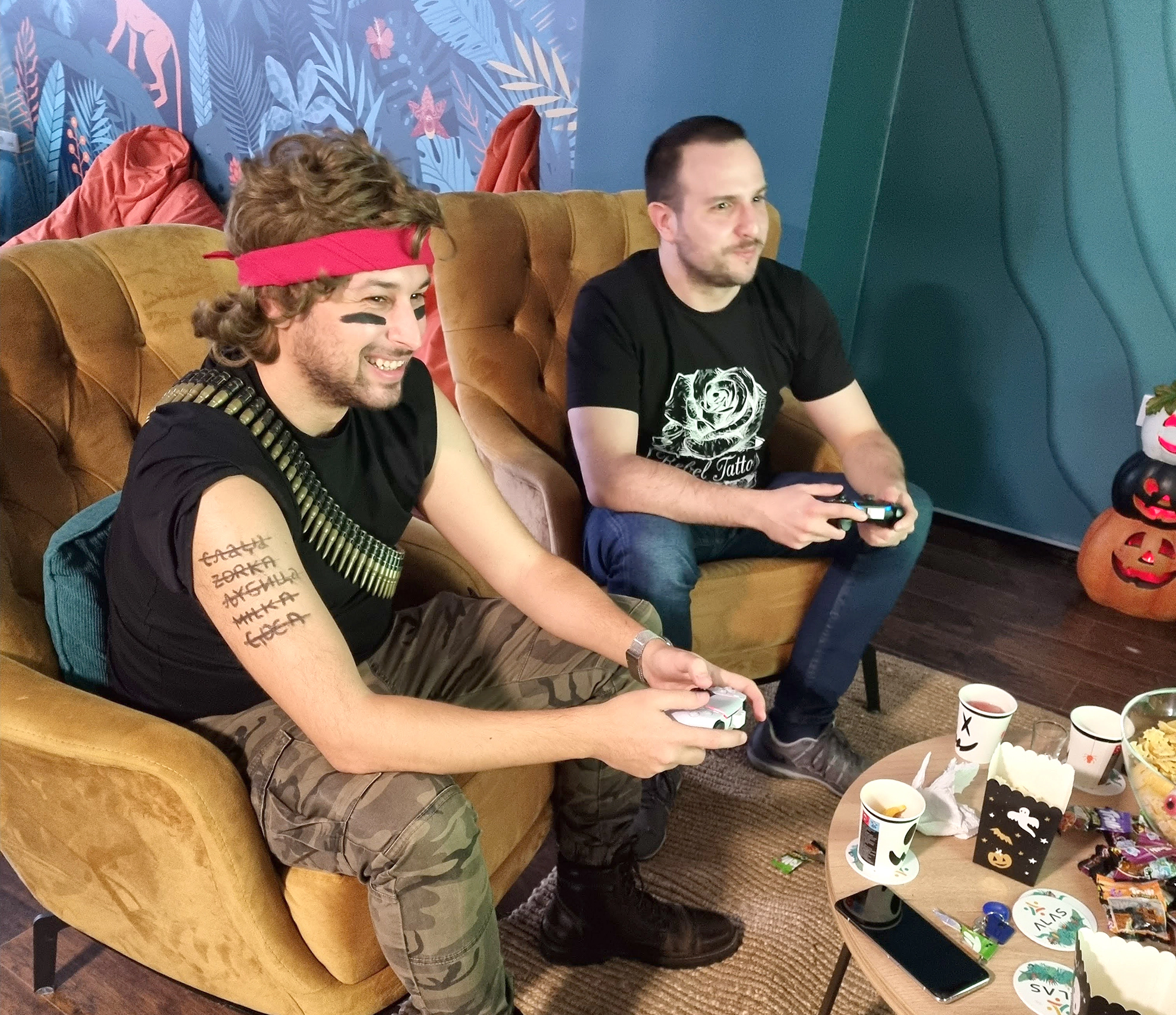 Two young men playing video games and eating snacks; one is in a Rambo halloween mask.