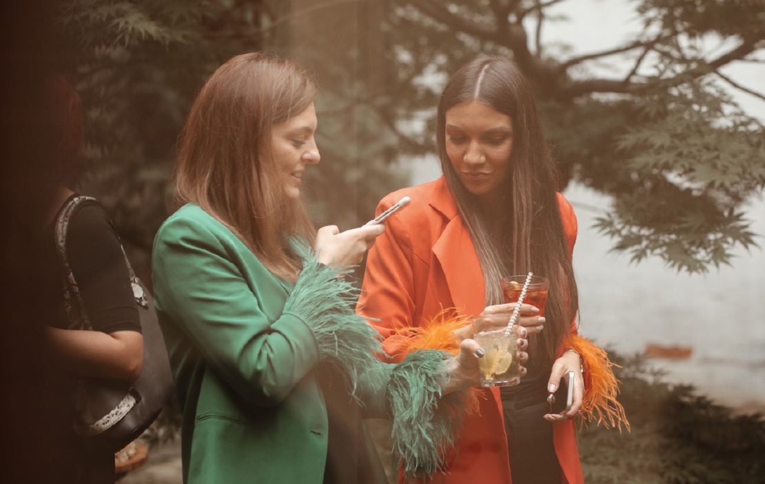 Two elegant young women taking photos of their coctails at an office party in the garden.